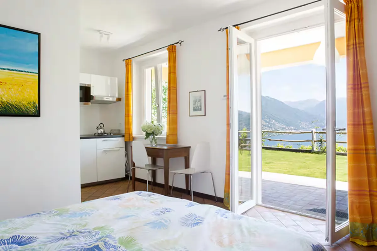 Interno del monolocale con il letto matrimoniale, l'angolo cottura bianco e la porta finestra aperta sul giardino con vista lago